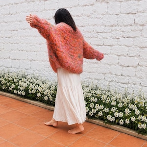 K&ouml;nnte beinhalten: Ein &uuml;bergro&szlig;er, flauschiger Langarmpullover in einer lebendigen Mischung aus korallenfarbenem, rosafarbenem und orangefarbenem Garn, kombiniert mit einem langen, flie&szlig;enden, cremewei&szlig;en Stufenrock. Der Pullover hat eine lockere Strickstruktur.