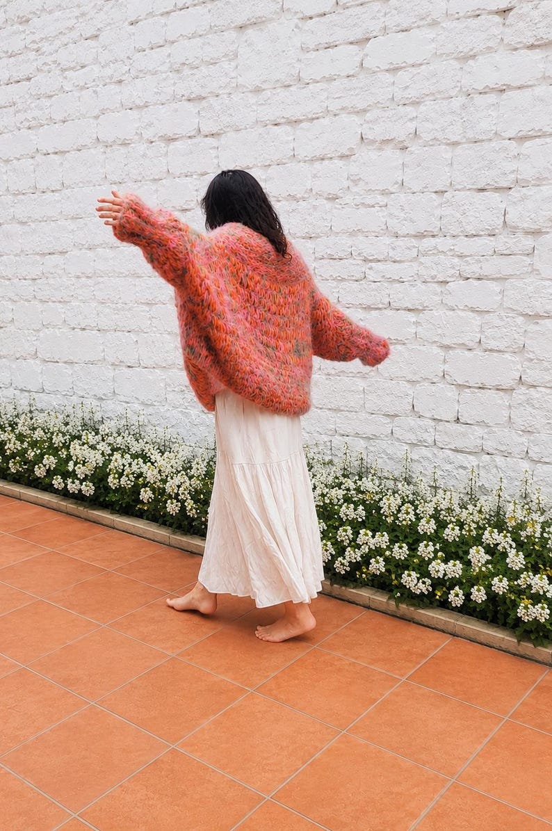 K&ouml;nnte beinhalten: Ein &uuml;bergro&szlig;er, flauschiger Langarmpullover in einer lebendigen Mischung aus korallenfarbenem, rosafarbenem und orangefarbenem Garn, kombiniert mit einem langen, flie&szlig;enden, cremewei&szlig;en Stufenrock. Der Pullover hat eine lockere Strickstruktur.