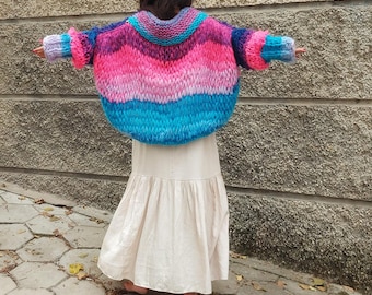 May include: A colorful, chunky knit sweater in shades of pink, blue, and purple. The sweater has a wide, rounded shape and long sleeves. The word "MOMA" is visible on the wall in the background. The person is wearing a long, cream-colored skirt.
