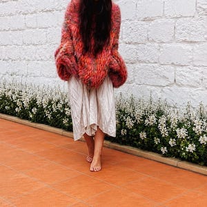K&ouml;nnte beinhalten: Ein &uuml;bergro&szlig;er, grob gestrickter Pullover mit einer leuchtenden Mischung aus rotem, orangenem und rosafarbenem Garn, kombiniert mit einem hellbeigen, wadenlangen Rock, der eine lockere, flie&szlig;ende Silhouette und eine leicht knittrige Textur aufweist.