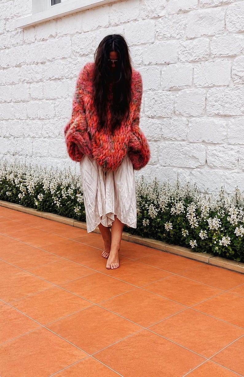 K&ouml;nnte beinhalten: Ein &uuml;bergro&szlig;er, grob gestrickter Pullover mit einer leuchtenden Mischung aus rotem, orangenem und rosafarbenem Garn, kombiniert mit einem hellbeigen, wadenlangen Rock, der eine lockere, flie&szlig;ende Silhouette und eine leicht knittrige Textur aufweist.