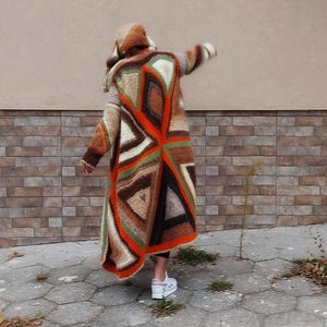 Puede incluir: Un c&aacute;rdigan largo de crochet marr&oacute;n, naranja, verde y blanco con un patr&oacute;n geom&eacute;trico. El c&aacute;rdigan tiene capucha y lo lleva puesto una persona de pie frente a una pared de ladrillo.