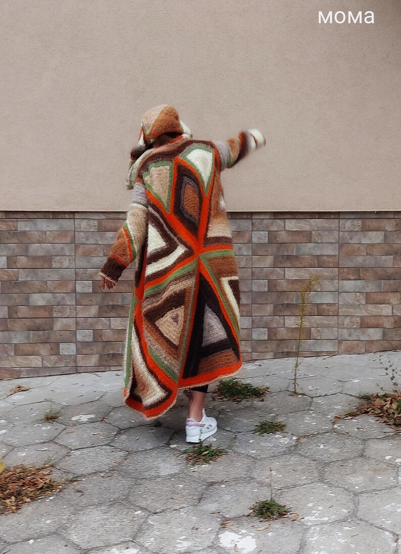 Puede incluir: Un c&aacute;rdigan largo de crochet marr&oacute;n, naranja, verde y blanco con un patr&oacute;n geom&eacute;trico. El c&aacute;rdigan tiene capucha y lo lleva puesto una persona de pie frente a una pared de ladrillo.