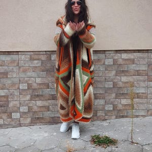 Puede incluir: Una mujer lleva una larga chaqueta de punto con capucha de color marr&oacute;n, naranja y verde. Est&aacute; de pie frente a una pared de ladrillo y lleva zapatillas blancas.