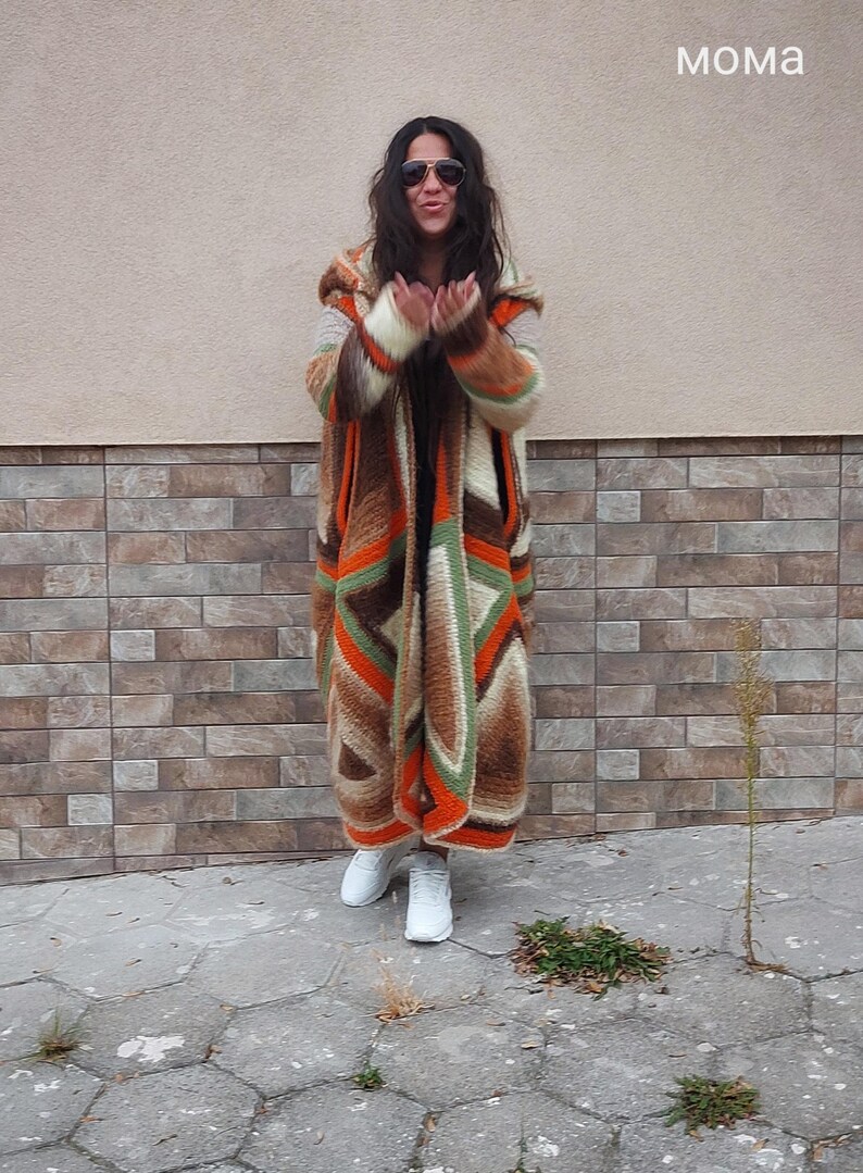 Puede incluir: Una mujer lleva una larga chaqueta de punto con capucha de color marr&oacute;n, naranja y verde. Est&aacute; de pie frente a una pared de ladrillo y lleva zapatillas blancas.