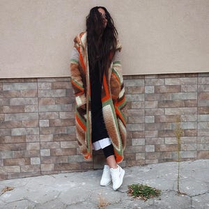 Puede incluir: Una mujer lleva una larga chaqueta de punto de ganchillo a rayas de colores con capucha. Lleva zapatillas blancas y est&aacute; de pie frente a una pared de ladrillo.