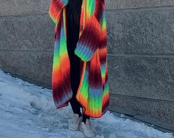 May include: A woman wearing a long, rainbow-colored knitted cardigan with a black beanie. The cardigan has a loose fit and is made of a chunky yarn. The woman is standing in front of a brick wall with snow on the ground.