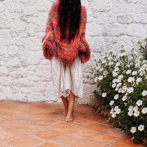 K&ouml;nnte beinhalten: Ein lang&auml;rmeliger, flauschiger Oversize-Pullover mit einem lebendigen, mehrfarbigen Strickmuster in Rosa-, Orange- und Rott&ouml;nen, kombiniert mit einem hellen, knielangen Rock mit gerafftem, flie&szlig;endem Saum und einer lockeren Passform.