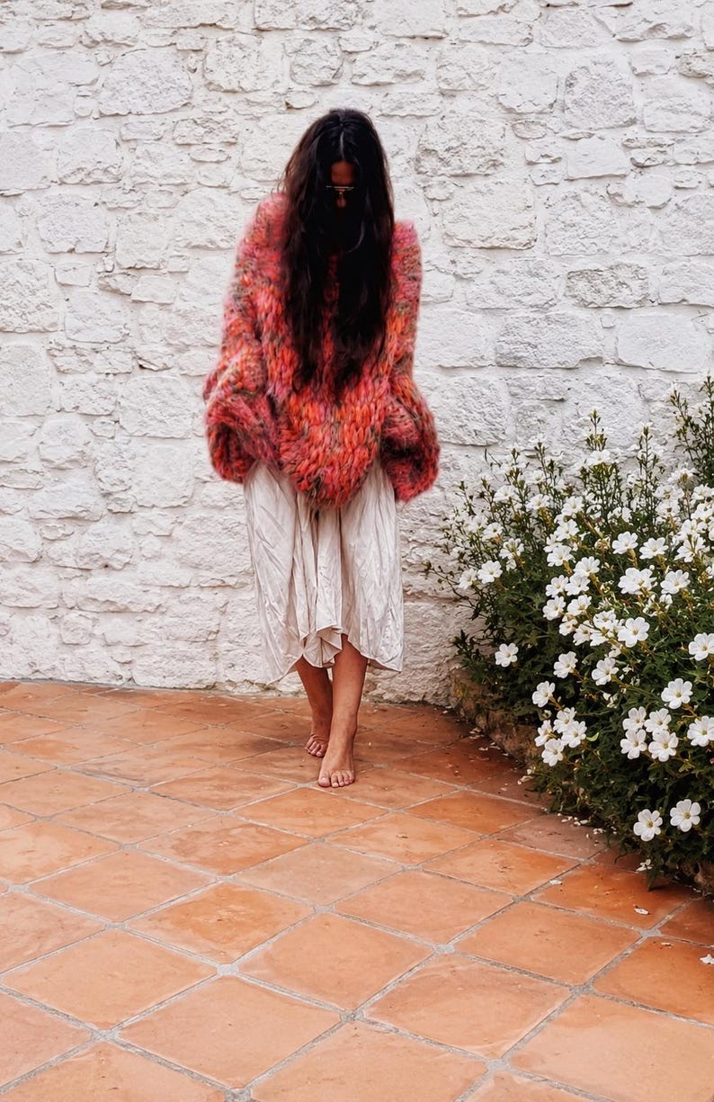 K&ouml;nnte beinhalten: Ein lang&auml;rmeliger, flauschiger Oversize-Pullover mit einem lebendigen, mehrfarbigen Strickmuster in Rosa-, Orange- und Rott&ouml;nen, kombiniert mit einem hellen, knielangen Rock mit gerafftem, flie&szlig;endem Saum und einer lockeren Passform.