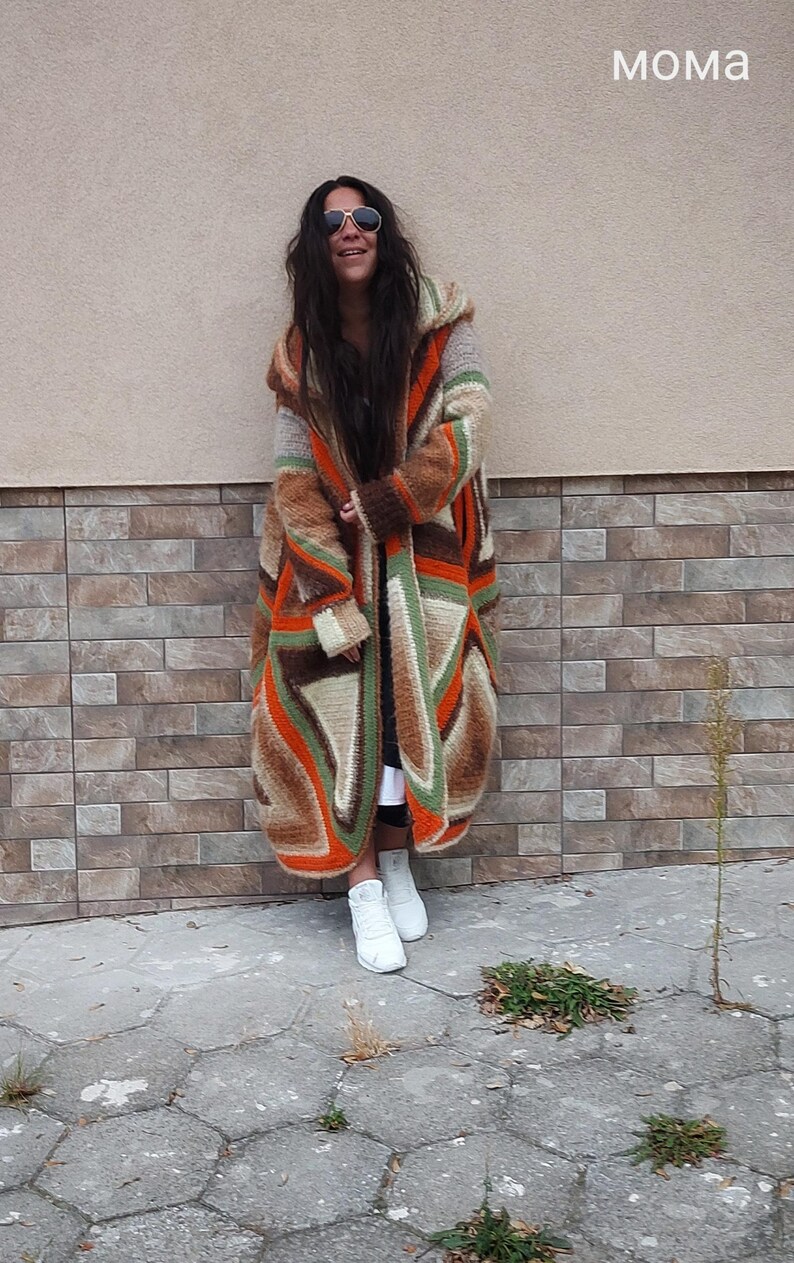 Puede incluir: Una mujer lleva una larga chaqueta de punto con capucha y estampado geom&eacute;trico de colores. Lleva zapatillas blancas y gafas de sol. La chaqueta est&aacute; hecha de un material suave y peludo.