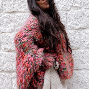 K&ouml;nnte beinhalten: Eine grob gestrickte, &uuml;bergro&szlig;e Strickjacke mit einer lebendigen, mehrfarbigen Garnmischung aus leuchtendem Pink, Orange und Gr&uuml;n. Das Kleidungsst&uuml;ck hat eine dicke, flauschige Textur mit langen &Auml;rmeln und wird &uuml;ber einem beigen, locker sitzenden Kleidungsst&uuml;ck getragen.