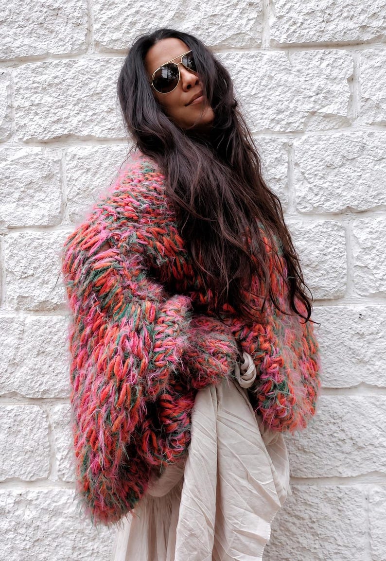 K&ouml;nnte beinhalten: Eine grob gestrickte, &uuml;bergro&szlig;e Strickjacke mit einer lebendigen, mehrfarbigen Garnmischung aus leuchtendem Pink, Orange und Gr&uuml;n. Das Kleidungsst&uuml;ck hat eine dicke, flauschige Textur mit langen &Auml;rmeln und wird &uuml;ber einem beigen, locker sitzenden Kleidungsst&uuml;ck getragen.