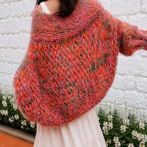 K&ouml;nnte beinhalten: Eine &uuml;bergro&szlig;e, grob gestrickte Strickjacke mit einer lebendigen Mischung aus korallenfarbenem, rosa, orangefarbenem und gr&uuml;nem Garn. Das Kleidungsst&uuml;ck hat ein lockeres, strukturiertes Gewebe mit weitem Kragen und langen &Auml;rmeln, getragen &uuml;ber einem wei&szlig;en Oberteil.