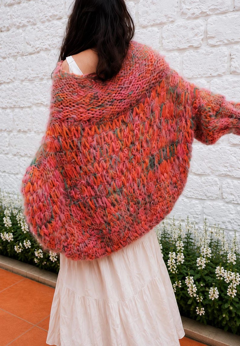 K&ouml;nnte beinhalten: Eine &uuml;bergro&szlig;e, grob gestrickte Strickjacke mit einer lebendigen Mischung aus korallenfarbenem, rosa, orangefarbenem und gr&uuml;nem Garn. Das Kleidungsst&uuml;ck hat ein lockeres, strukturiertes Gewebe mit weitem Kragen und langen &Auml;rmeln, getragen &uuml;ber einem wei&szlig;en Oberteil.