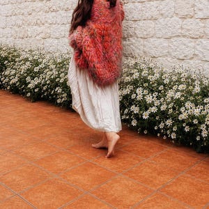 K&ouml;nnte beinhalten: Eine lang&auml;rmelige, grob gestrickte Strickjacke in verschiedenen Rosa-, Orange- und Rott&ouml;nen, getragen &uuml;ber einem schlichten, bodenlangen, cremewei&szlig;en Kleid. Die Strickjacke zeichnet sich durch ein lockeres, strukturiertes Gewebe aus.