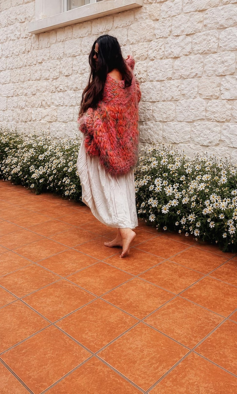 K&ouml;nnte beinhalten: Eine lang&auml;rmelige, grob gestrickte Strickjacke in verschiedenen Rosa-, Orange- und Rott&ouml;nen, getragen &uuml;ber einem schlichten, bodenlangen, cremewei&szlig;en Kleid. Die Strickjacke zeichnet sich durch ein lockeres, strukturiertes Gewebe aus.