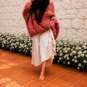 K&ouml;nnte beinhalten: Ein &uuml;bergro&szlig;er, grob gestrickter Pullover in einer bunten Mischung aus leuchtend rosa, orangefarbenem und gelbem Garn, kombiniert mit einem cremefarbenen, wadenlangen Rock mit subtiler Knitterstruktur und asymmetrischem Saum.