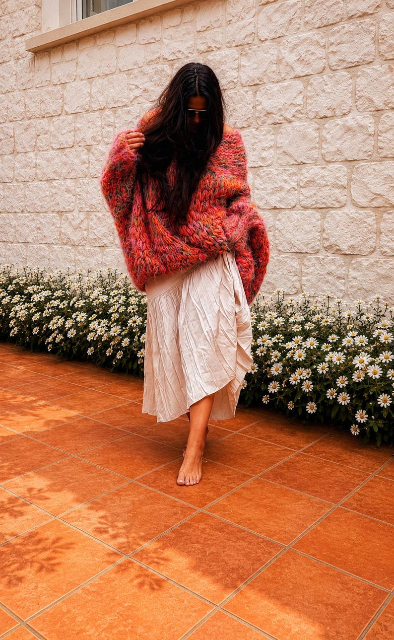 K&ouml;nnte beinhalten: Ein &uuml;bergro&szlig;er, grob gestrickter Pullover in einer bunten Mischung aus leuchtend rosa, orangefarbenem und gelbem Garn, kombiniert mit einem cremefarbenen, wadenlangen Rock mit subtiler Knitterstruktur und asymmetrischem Saum.