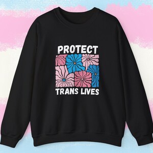 Pode incluir: Camisola preta com o texto branco "PROTECT TRANS LIVES". O texto está acima de um desenho quadrado de flores rosa e azuis. O fundo é um gradiente azul claro, rosa e branco.