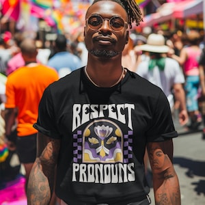 May include: A person wearing a black t-shirt with the words "Respect Pronouns" in a retro font. The shirt has a floral design in purple and yellow. The background shows a crowd and rainbow flags.