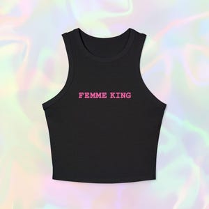 Op de afbeelding: Zwarte crop top met de roze tekst "FEMME KING" op de voorkant. De top is mouwloos en heeft een ronde hals. De achtergrond is een vage, iriserende patroon van roze, paars en groen.