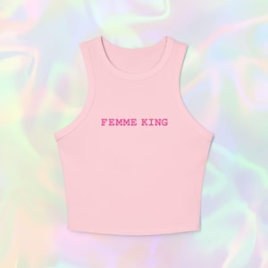 Op de afbeelding: Een lichtroze tanktop met de tekst "FEMME KING" in een bijpassende roze kleur. De top heeft een racerback-ontwerp en een ronde hals. De achtergrond is een holografisch, iriserend patroon.
