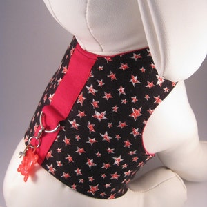 Może przedstawiać: Czarno-czerwona szelka dla psa z białym wzorem w gwiazdki. Szelka ma czerwony pasek i srebrny pierścień do mocowania smyczy. Do pierścienia przymocowany jest czerwony plastikowy zabawka.