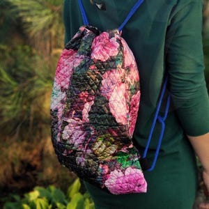 Può includere: Una borsa a cordoncino floreale con un design trapuntato. La borsa è rosa, verde e nera con un cordoncino blu.