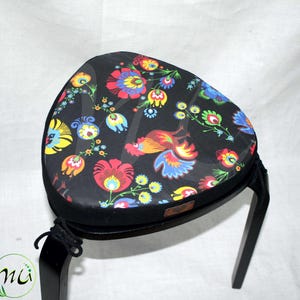 May include: A black wooden stool with three legs and a round seat. The seat is covered in black fabric with a colorful floral pattern featuring roosters and flowers.