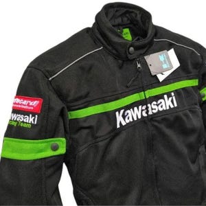 Puede incluir: Chaqueta de moto negra con detalles verdes y blancos. Presenta el logotipo de Kawasaki en el pecho y una franja verde. Incluye un pequeño parche rojo con texto blanco y una etiqueta.