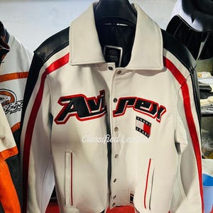 Könnte beinhalten: Weiße Lederjacke mit schwarzen und roten Akzenten. Sie trägt den Schriftzug "Avirex" in roter und schwarzer Schrift sowie einen Tommy Jeans-Patch. Kragen, Bündchen und Saum sind gerippt.