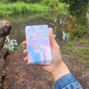 Può includere: Un mazzo di carte dei tarocchi con un design celeste colorato è tenuto in mano. Le carte presentano un mix di colori pastello, tra cui blu, viola e rosa, con forme geometriche. Lo sfondo mostra un ambiente naturale all'aperto con uno specchio d'acqua.