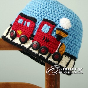 May include: A blue crocheted hat with a red, yellow, and black train design. The train has black buttons for wheels and a white pom-pom on top.