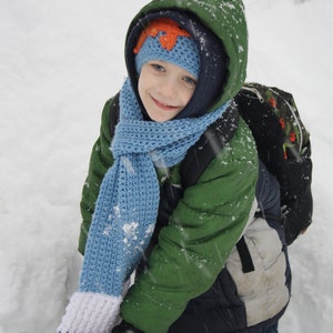 May include: A young child wearing a green winter jacket, a blue knitted scarf, and a blue and orange knitted hat stands in the snow. The child is smiling and looking at the camera.