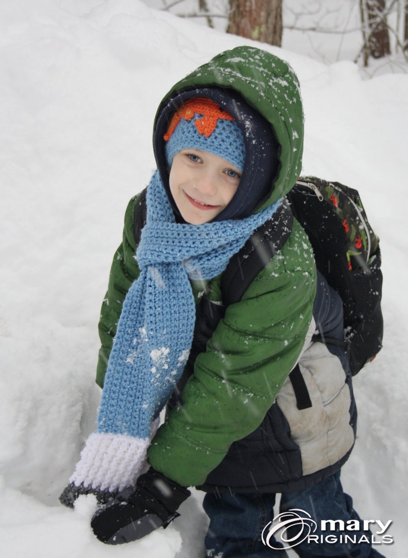 May include: A young child wearing a green winter jacket, a blue knitted scarf, and a blue and orange knitted hat stands in the snow. The child is smiling and looking at the camera.