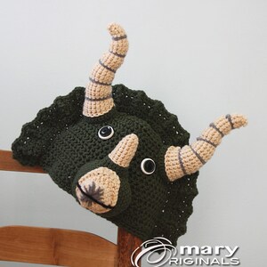 Puede incluir: Sombrero de triceratops de crochet verde con cuernos beige y ojos negros. El sombrero est&aacute; sentado en una silla de madera.