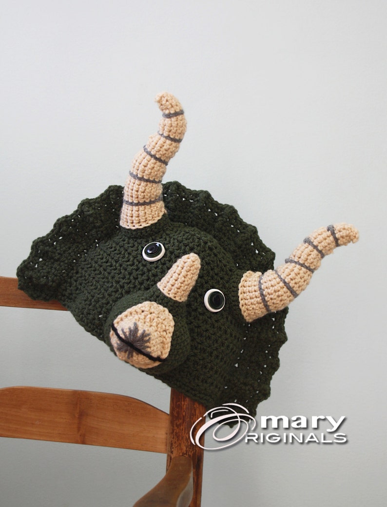 Puede incluir: Sombrero de triceratops de crochet verde con cuernos beige y ojos negros. El sombrero est&aacute; sentado en una silla de madera.
