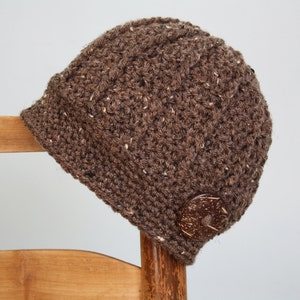 May include: A brown crocheted hat with a small brim and a decorative wooden button. The hat is made of a textured yarn with flecks of white. The hat is resting on a wooden chair.