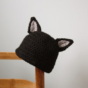 May include: A black crocheted hat with gray crocheted ears. The hat is sitting on a wooden chair.