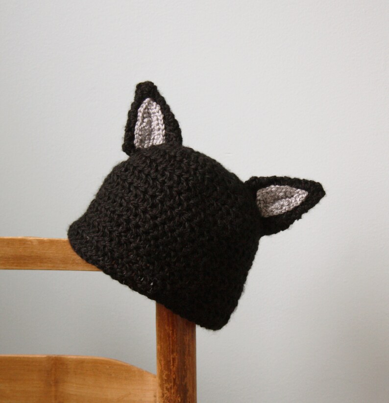 May include: A black crocheted hat with gray crocheted ears. The hat is sitting on a wooden chair.