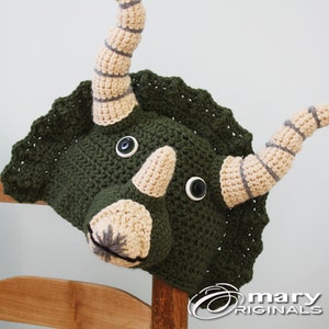 Puede incluir: Sombrero de triceratops de crochet verde con cuernos y ojos beige. El sombrero tiene un borde festoneado y est&aacute; sentado en una silla de madera.
