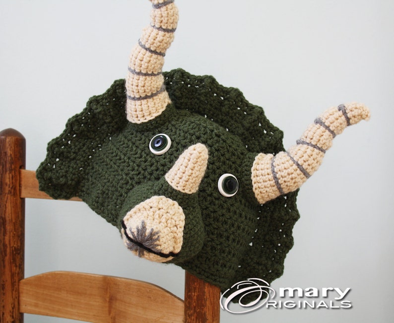 Puede incluir: Sombrero de triceratops de crochet verde con cuernos y ojos beige. El sombrero tiene un borde festoneado y est&aacute; sentado en una silla de madera.