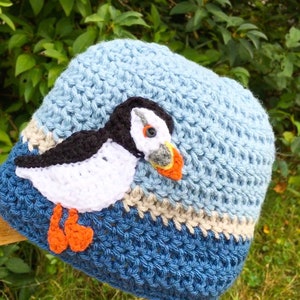 May include: A blue and white crocheted hat with a puffn bird design. The hat has a light blue top, a white stripe, a beige stripe, and a dark blue bottom.
