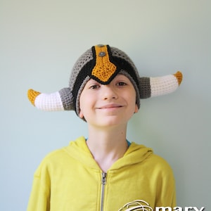 May include: A child wearing a gray, black, and yellow crocheted viking hat with horns. The hat has a button on top. The child is smiling and wearing a yellow hoodie.