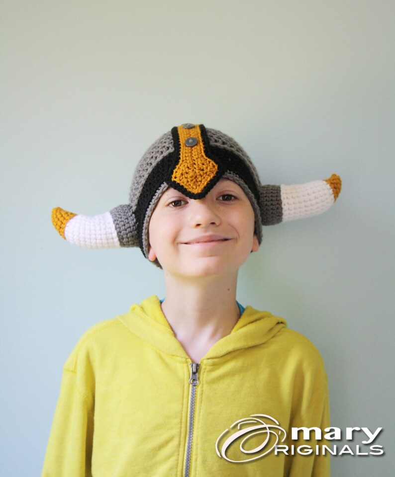 May include: A child wearing a gray, black, and yellow crocheted viking hat with horns. The hat has a button on top. The child is smiling and wearing a yellow hoodie.