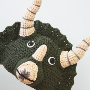 Puede incluir: Un gorro de triceratops de crochet verde con cuernos beige y rayas grises. El gorro tiene dos botones negros grandes para los ojos.