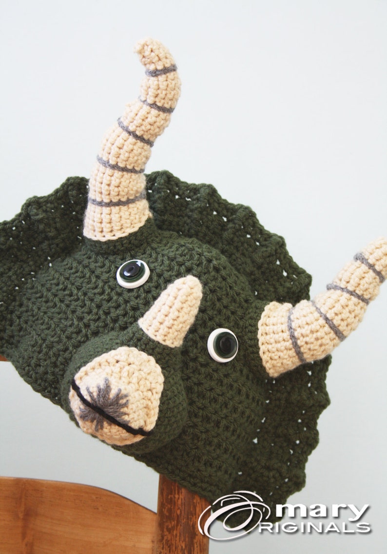 Puede incluir: Un gorro de triceratops de crochet verde con cuernos beige y rayas grises. El gorro tiene dos botones negros grandes para los ojos.