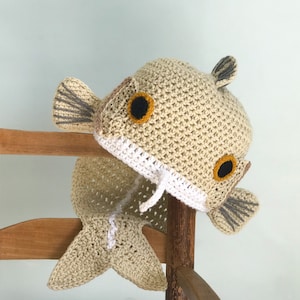 Könnte beinhalten: Eine gehäkelte beige und weiße Fischmütze mit einem Schwanz. Die Mütze hat schwarze und gelbe Augen und eine graue Flosse.