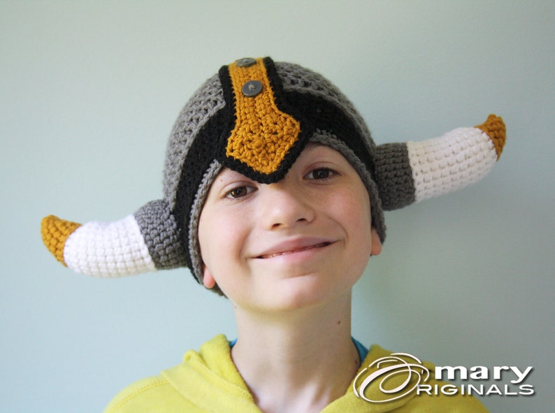 May include: A child wearing a gray, black, and yellow crocheted viking hat with white horns.