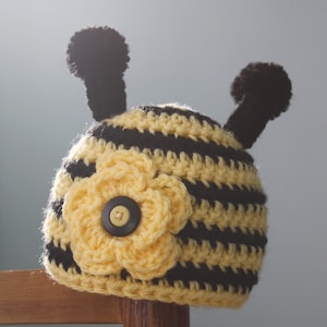 Può includere: Un cappello da ape a righe gialle e nere lavorato a uncinetto con un fiore giallo e un bottone nero. Il cappello ha due pompon neri in cima.